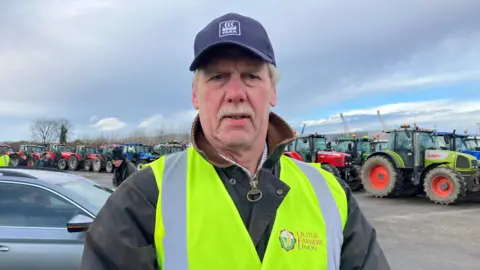 A man, with grey hair and a grey moustache, wearing a jacket and hi-vis vest. He is also wearing a navy cap. There are tractors in the background. 