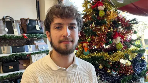 John Devine/BBC Nick Lambert who has mid brown hair in a fluffy fringe on his forehead and a short dark beard. He is wearing a cream jumper over a white polo-shirt. He is standing his father's shop with a shelves holding purses and bags behind him on the left and a Christmas tree covered in tinsel on his right. 