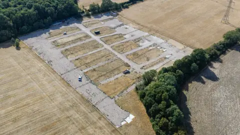 Supplied An aerial shot of the site, which is now clear of mobile homes and caravans, although two small vehicles can be seen. The hardstanding remains and the grass around it is now brown due to the drought. Green trees are at the edges.