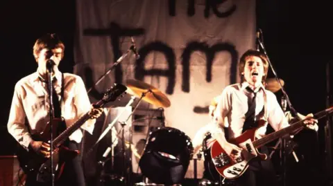 Getty Images An old image of two people performing on a stage with guitars. In the background is a hand painted banner with "The Jam" on it.