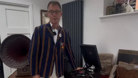 Mr Wareham wears a blue and yellow striped blazer and brown dicky-bow tie.   He stands next to one turn table, with the horn gramophone behind him.  There are vintage records on the table.