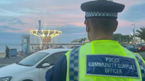 Rear view of a PCSO wearing a high-vis vest and a hat with Weymouth beach and a funfair in the background