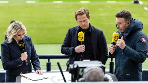 SNS Three pundits, a woman and two men, stand facing a TV camera holding yellow and black microphones which read "PS". They are on a balcony which looks on to the pitch for Hampden Park