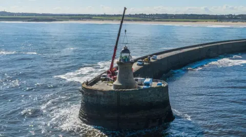 Dave Bell / Port of Tyne A red crane placing the new dome on relatively short, brick lighthouse. It sits on the circular end of a brick pier which arcs back to the shoreline. The lighthouse is surrounded by white vans and the coast is in the distance.