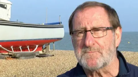 Andy Roberts is standing on a shingle beach in Kent with a boat over his right hand shoulder.