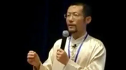 CPS A still taken from a video of a presentation by Hongchi Xiao on Paida Lajin therapy. Xiao wears a long white jacket  and is holding a microphone.