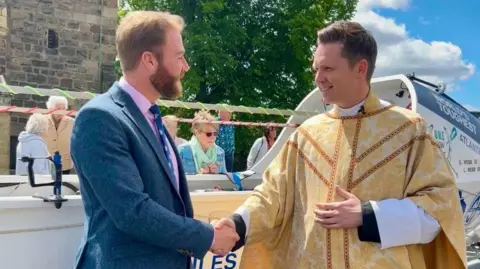 Louis Webb A man dressed in a blue suit is stood in front of a large sailing boat shaking hands with a vicar
