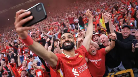 A grinning Mohamed Salah, in red club kit, takes a selfie with a mobile phone in front of excited Liverpool supporters