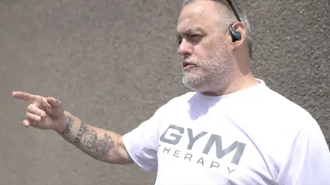A man with grey hair and beard us pointing to the right, he is wearing a white t shirt. The background is a gravel wall. 