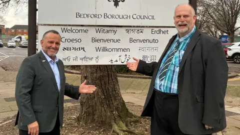 Bedford Borough Council Two men smiling in front of a sign that says "Bedford Borough Council".