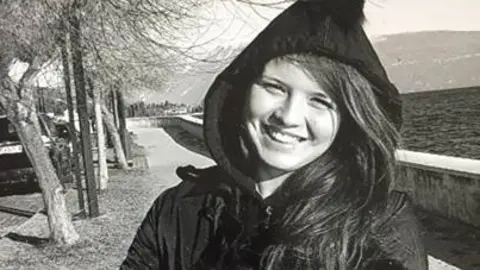 A black and white photo of the late Natasha Melendez.  She has long, straight hair and is smiling at the camera.  She is wearing a dark, hooded coat.  She is standing on a tree-lined path with a wall and a large body of water behind her. 