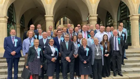 Laura Coffey/BBC Men and women wearing suits, standing on steps outside the Guildhall in Northampton. Many of them are wearing light blue rosettes and council lanyards.