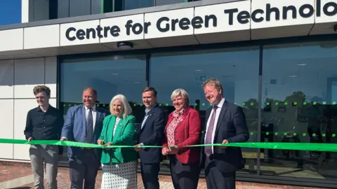 A line of dignitaries at the opening of the centre. They are standing behind a green ribbon which a woman in a green jacket is about to cut with a pair of scissors.