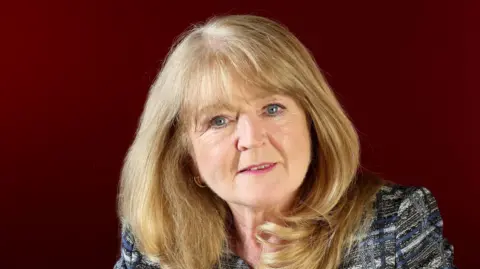 Equality Commission for Northern Ireland Geraldine McGahey. She has long blonde hair with a fringe, blue eyes, wearing a dark grey blazer. Behind her is a dark red background.