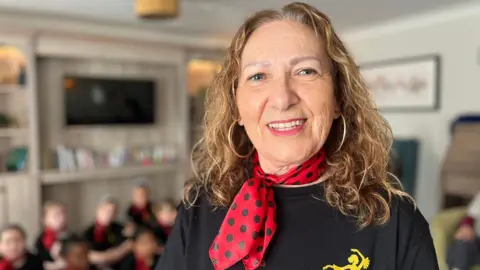 George Carden/BBC Josefa looks at the camera with blurred children in the background. They are all wearing a black t-shirt with a woman dancing in the top left hand corner as well as a red and black polka-dot scarf. She has blonde, curly hair and hoop earrings. 