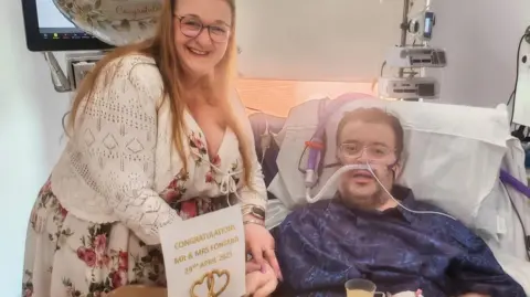 A man with a high flow nasal cannula and wearing a navy shirt and clear-framed glasses lying in a hospital bed, on his left a lady in a white dress with flowers on it with a white cardigan over wearing glasses holds his hand. In front of them is a sign that reads: 'Congratulations Mr & Mrs Fontana 29th April 2025', a balloon is behind the lady's head.