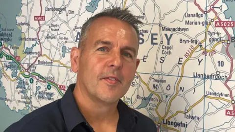 Mike Williams is photographed standing in front of a map of Anglesey. He has short grey hair and is wearing a black shirt. 
