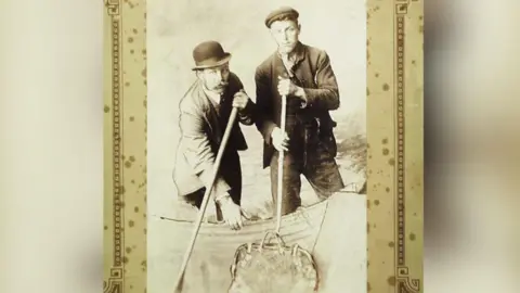 Copyright: Reading Museum A black and white sepia style posed photograph of two men in a boat holding barge poles. One of the poles has a carpet bag hooked on the end. They are wearing victorian attire. One has a bowler hat on, the other has a flat cap. The image has an aged, patterned cardboard frame.
