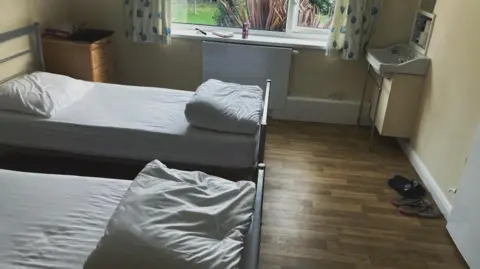 Shantel Morris A photo of a wooden floored room with two single beds and a window overlooking a grass lawn. The beds have white sheets on them and a folded white duvet at the foot of the bed and pillows at the top.