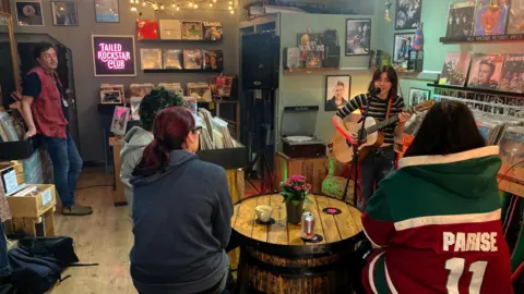 BBC A guitarist is sitting on a stool singing to coffee shop customers