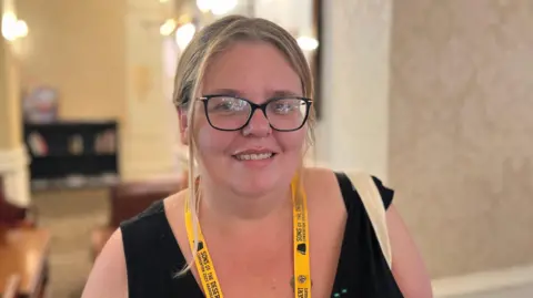 A woman with tied back blonde hair, wearing a black top, yellow lanyard and black glasses, smiles at the camera.