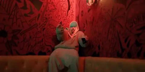 Justin Bieber Justin Bieber wearing a hooded top and beanie hat and holding a drink, sits in a pub with an illustrated wall in the background