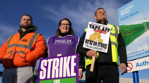 Two men and a woman stand on a picket line with a large Scottish Water billboard - featuring a glass of water against a scenic backdrop - to their right.