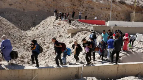 Reuters Dozens of people carrying their childen and belongings walk through the cratered road to reach the Masnaa crossing taking them into Syria on 4 October 2024.