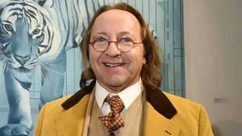 Bill Pallot smiles in front of a painting at a gallery wearing round-rimmed glasses and a three-piece yellow suit.