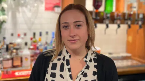 Emily McNeil, a 25-year-old white woman with short blonde and brown hair. She is wearing a cream spotted shirt with a black cardigan