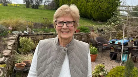 A woman with short light brown hair and dark rimmed glasses stands at the bottom of a garden in a stoned courtyard. There are several pots and plants behind her and a patio table and chairs. She is wearing a white longsleeved top and a beige gilet.