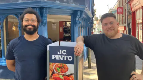 Owen Sennitt/BBC A man with glasses and a blue top and another man with a dark top look towards the camera and smile in front of a café named JC Pinto in Norwich, which is painted blue, with a narrow street visible behind them.