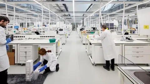 A white laboratory-type building with white work stations where people in white lab coats are working. The work stations have drawers underneath them. A glass roof is held up by white poles.