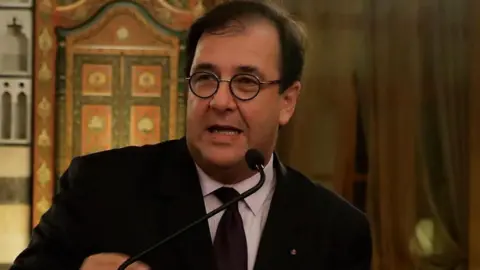 Bruno Foucher in a black suit and tie and a white shirt speaks to a microphone