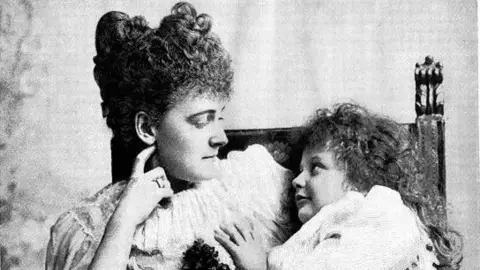 Getty Images A black-and-white image of a mother with a child on her knee. The woman, on the left, has curly hair piled high on her head and is wearing a dress with frills at the collar. She has her hand touching her face as she gazes at the child. The girl, who has long curly dark hair, is looking at her mother and wearing a white gown.