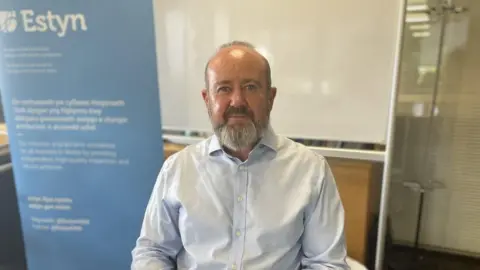 Owen Evans sitting in the class in front of a white board and blue sign which says Estyn in white writing