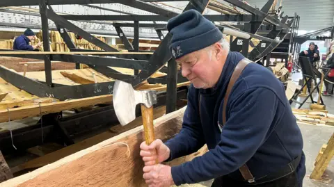 Stuart Howells/BBC A volunteer holds an axe next to a plank of wood he aims to chop for the project. He  wears a navy jumper as well as a navy beanie hat.