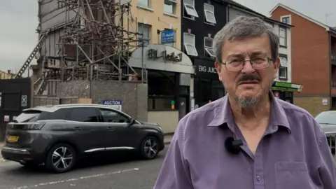 Giles du Boulay, wearing a purple shirt, surrounded by scaffolding, with cars passing in the foreground and shops and flats in the background.