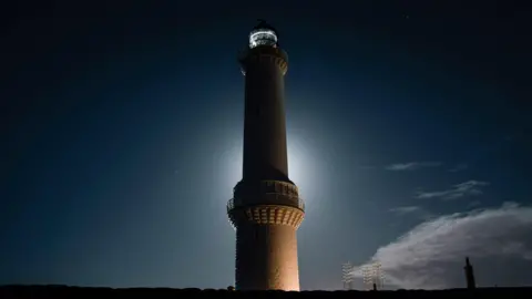 Andrew Gilchrist A tall lighthouse rises against a dark night sky, its illuminated tower glowing softly with a halo of light and faint stars overhead.
