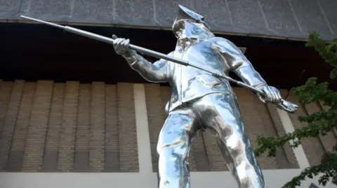 Stoke-on-Trent City Council A metal statue showing a life-sized man dressed in steelworkers' uniform and holding a long pole with both hands. There is a light-coloured brick building in the background.