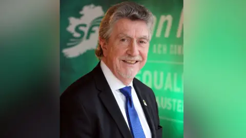 Getty Images Brady is pictured smiling at the camera for an election campaign poster. He has short greying hair, a grey goatee and moustashe. He is wearing a black suit with a blue tie and white shirt. The background says Sinn Féin