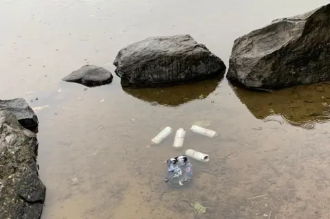 Emily Crichton Four beer cans and plastic bottles floating in a loch beside rocks