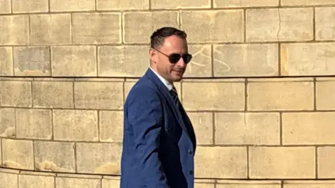 Jason Zadrozny in a blue suit and wearing sunglasses walking next to a pale stone wall
