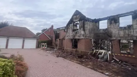 A large two-storey brick house severely damaged by fire, with the roof completely destroyed and upper walls blackened by smoke and scorch marks. Burnt window openings and debris surround the building, while caution tape cordons off the front. A detached three-car garage with white doors stands intact beside a paved driveway under an overcast sky.