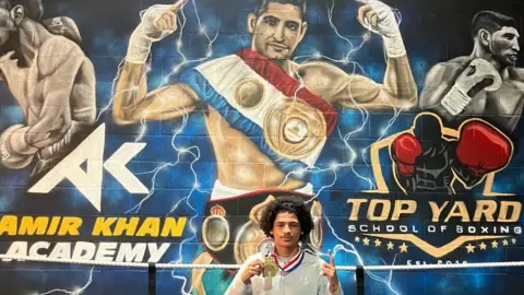 Shariqua Ahmed/ BBC Ismael wearing a grey track suit and a gold medal around his neck standing in front a Amir Khan's Mural on the wall facing the boxing ring 
