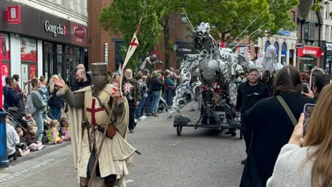 Phil Harrison/BBC A man dressed as St George and a large mechanical dragon head through crowds down a high street 