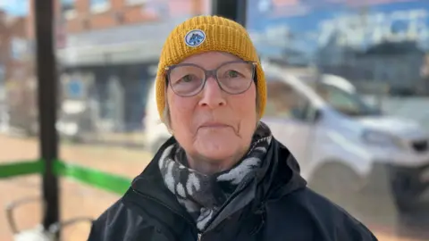 Susan Cannell, a white woman in her 60s, wearing a black coat and a printed black and white scarf underneath, and a yellow woolly hat. She is sitting in a bus stop shelter.