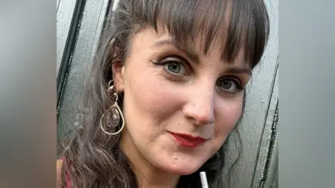 Woman with long brown hair smiles at camera. She has a gold earring and wears red lipstick