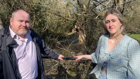 A shaven-headed man stands to the left of the picture in a purple shirt and black rain jacket. He holds a black guard rail with his left hand, next to a blonde woman in a blue checked blouse. Behind them is a stream with trees growing throughout.