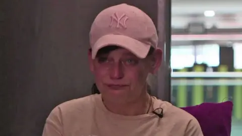 BBC Collette, wearing a pink t-shirt and pink baseball cap, looks into the camera with tears in her eyes 